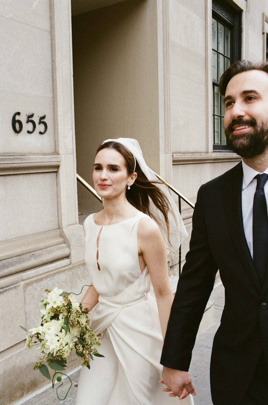 Bride in a white wedding dress holding a bouquet, walking with a bearded man in a suit as they exit a stone building, holding hands.