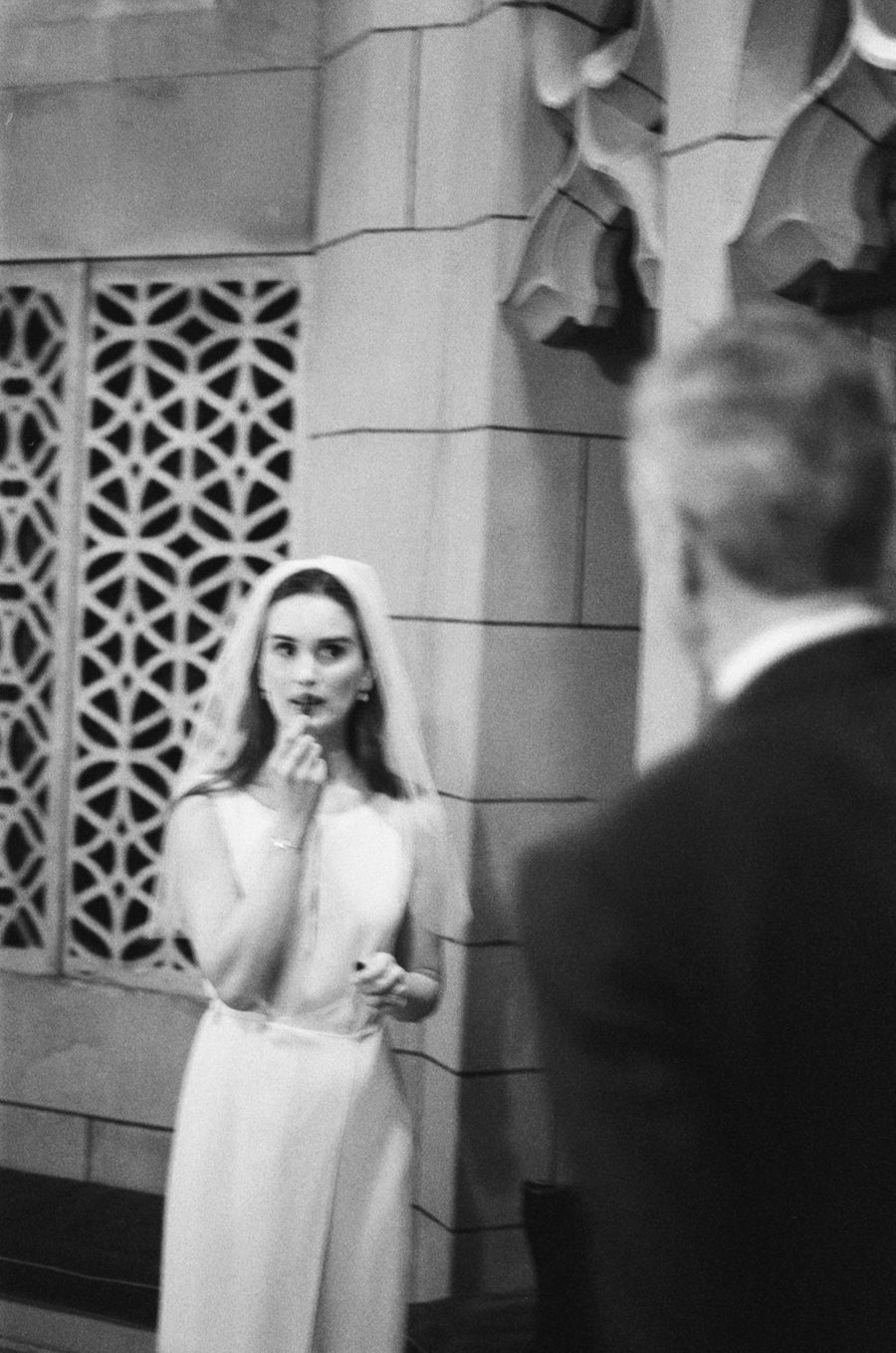 Bride in a white wedding dress and veil applying lipstick beside a patterned stone wall, man blurred in the foreground