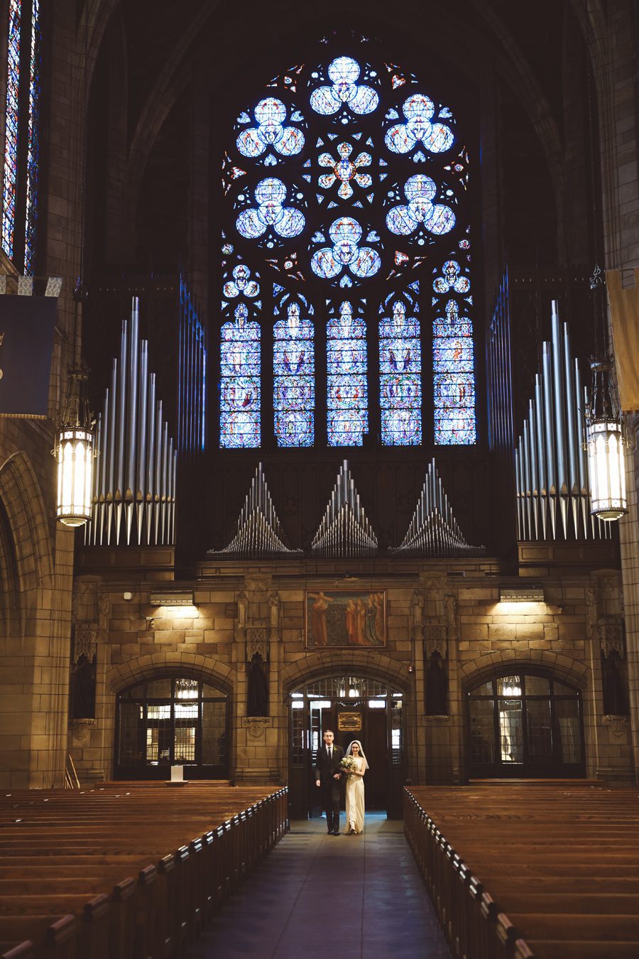 Bride in a white gown and groom in a dark suit walk hand in hand down the church aisle beneath a towering stained-glass window and organ pipes behind them.