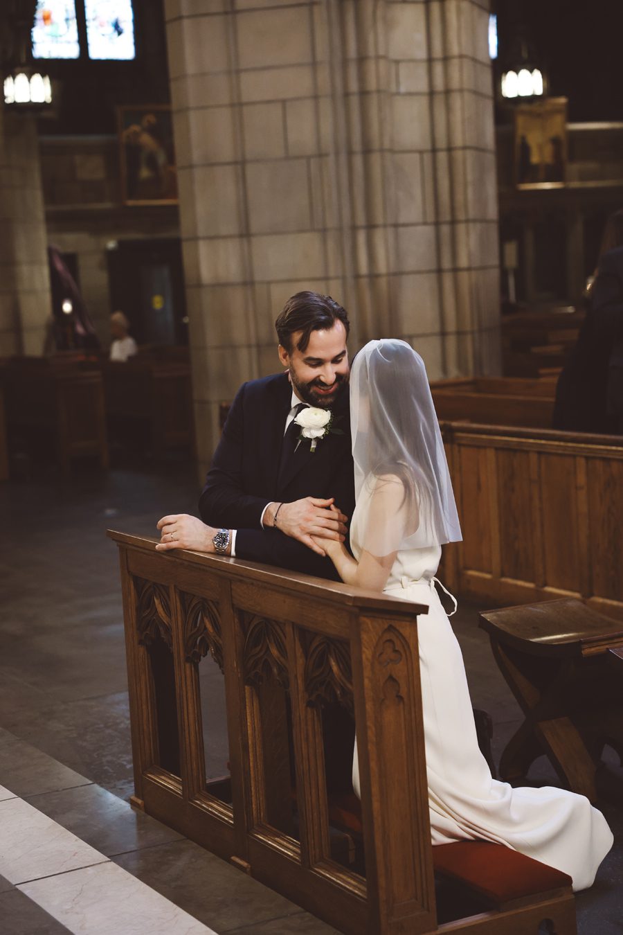 A bride and groom kneel at a wooden church pew, sharing a tender moment with the groom's hands clasped and the bride wearing a veil and white dress.