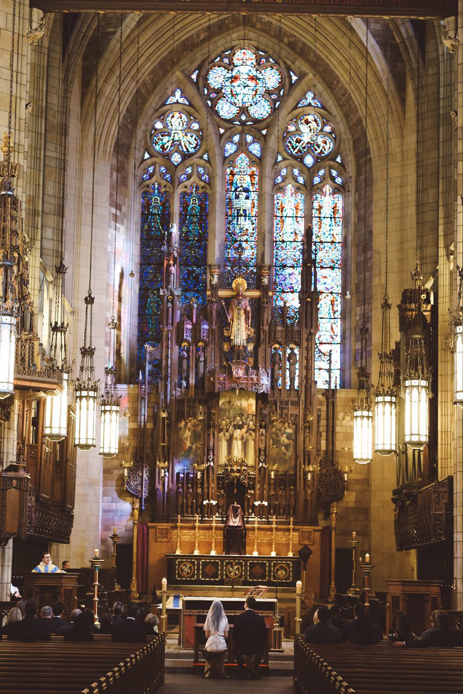 Bride and groom kneel at the altar in a Gothic church, with colorful stained-glass windows behind them.
