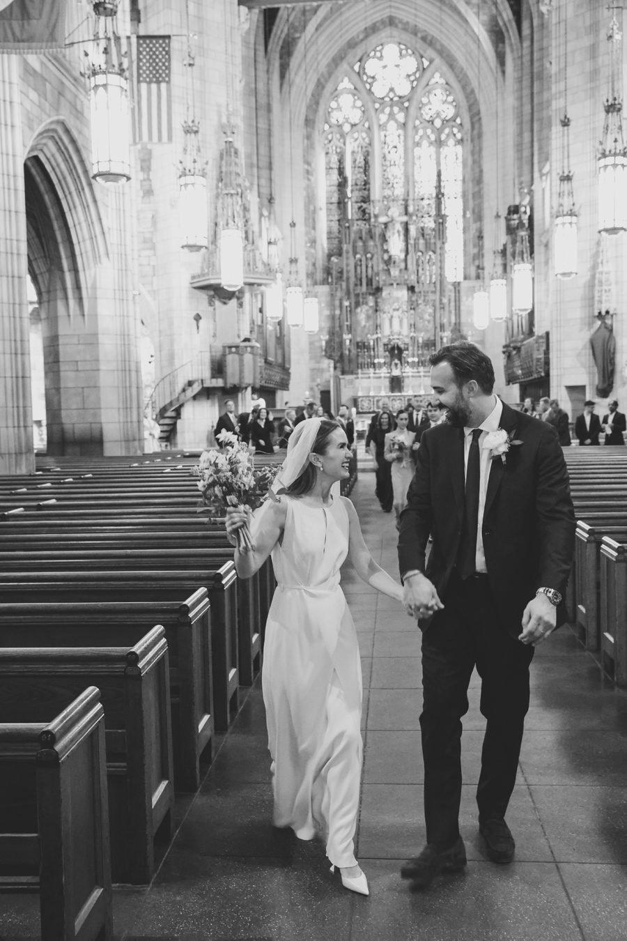 Bride and groom walking hand in hand down a church aisle, the bride holding a bouquet and smiling at him.
