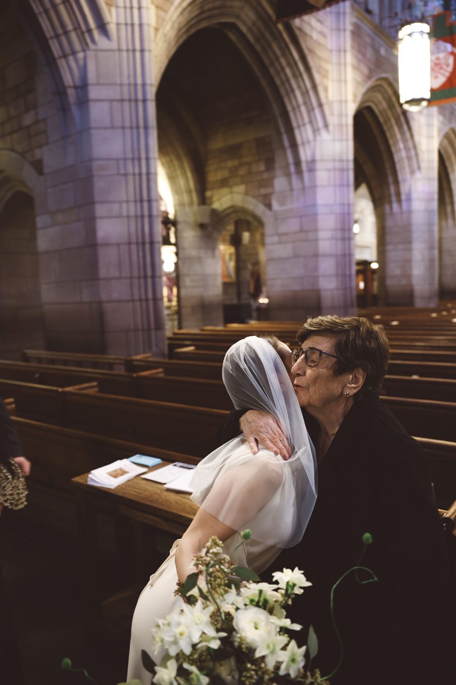 Bride wearing a veil hugs and kisses an older woman inside a stone church, bouquet in foreground and wooden pews in the background