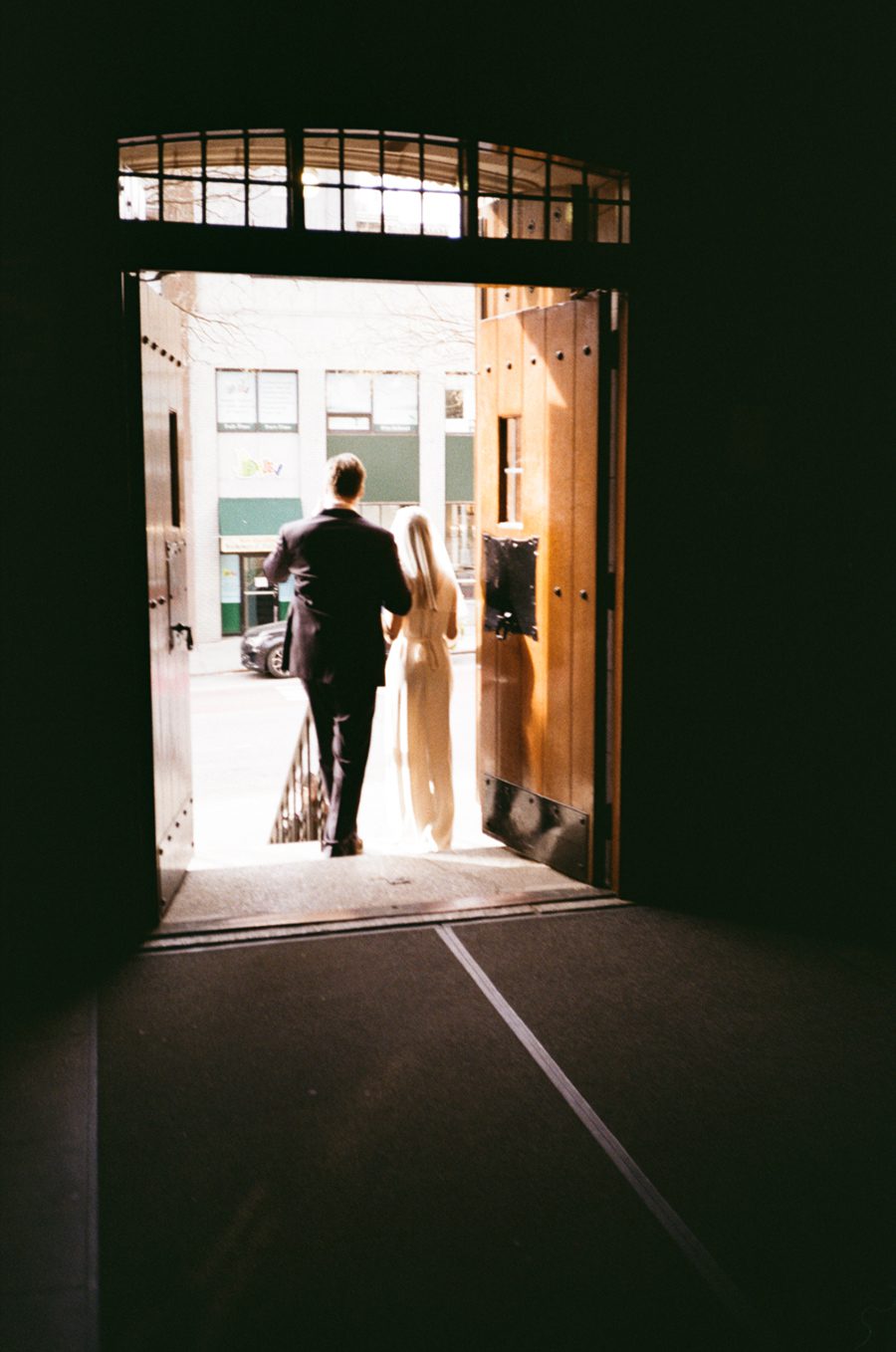 Couple in formal attire exit a dark interior through an open wooden door to a sunlit street outside.