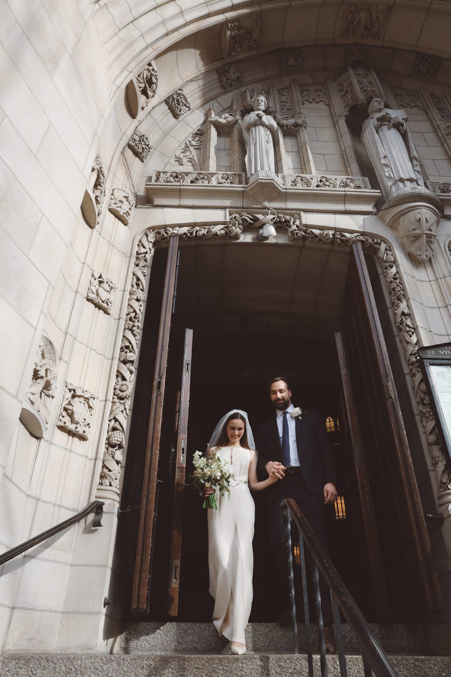 Bride and groom exit an ornate stone church doorway; she wears a white jumpsuit with a veil and bouquet while he in a dark suit helps her down the steps.