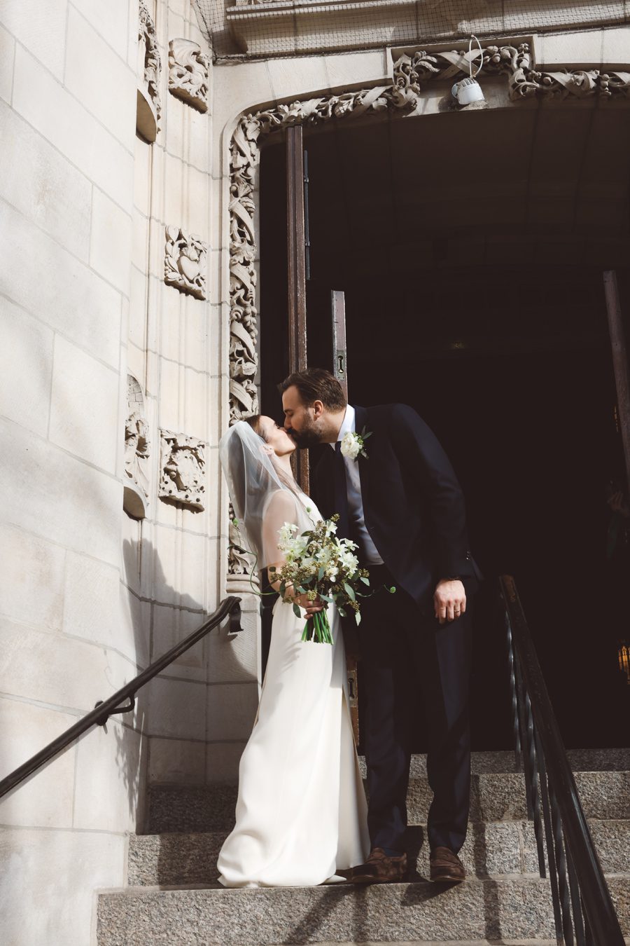 Bride in a white gown and veil kisses a groom in a dark suit on stone church steps, bouquet in hand.