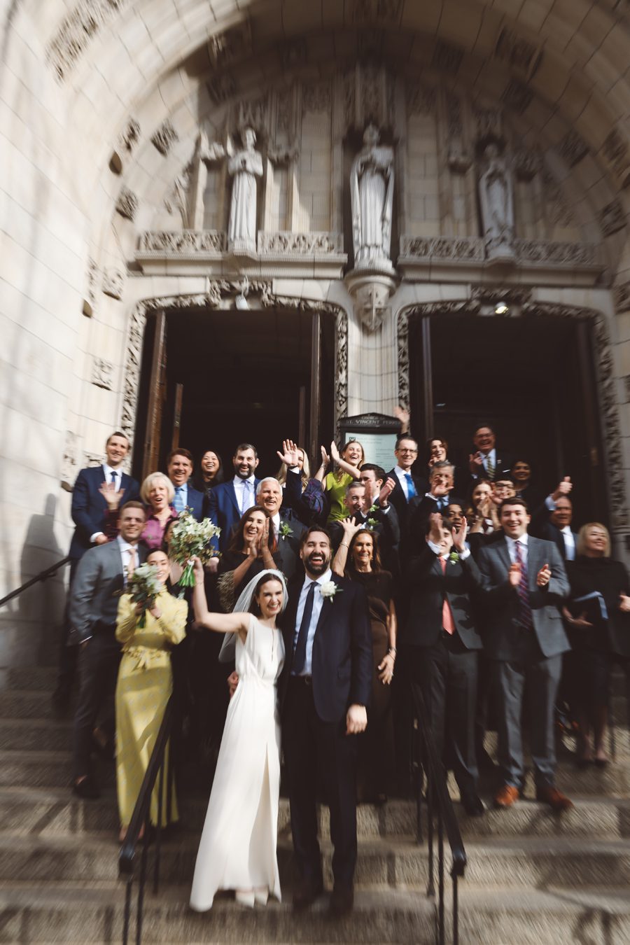 Bride and groom stand center on stone stairs, waving, surrounded by smiling wedding guests outside a church.
