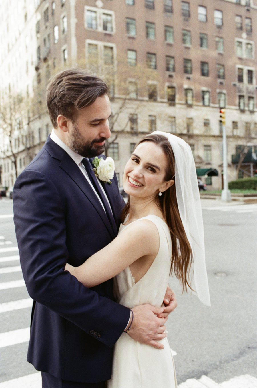 Bride and groom embrace on a city street; she smiles in a white dress with veil, he in a navy suit with a boutonniere.