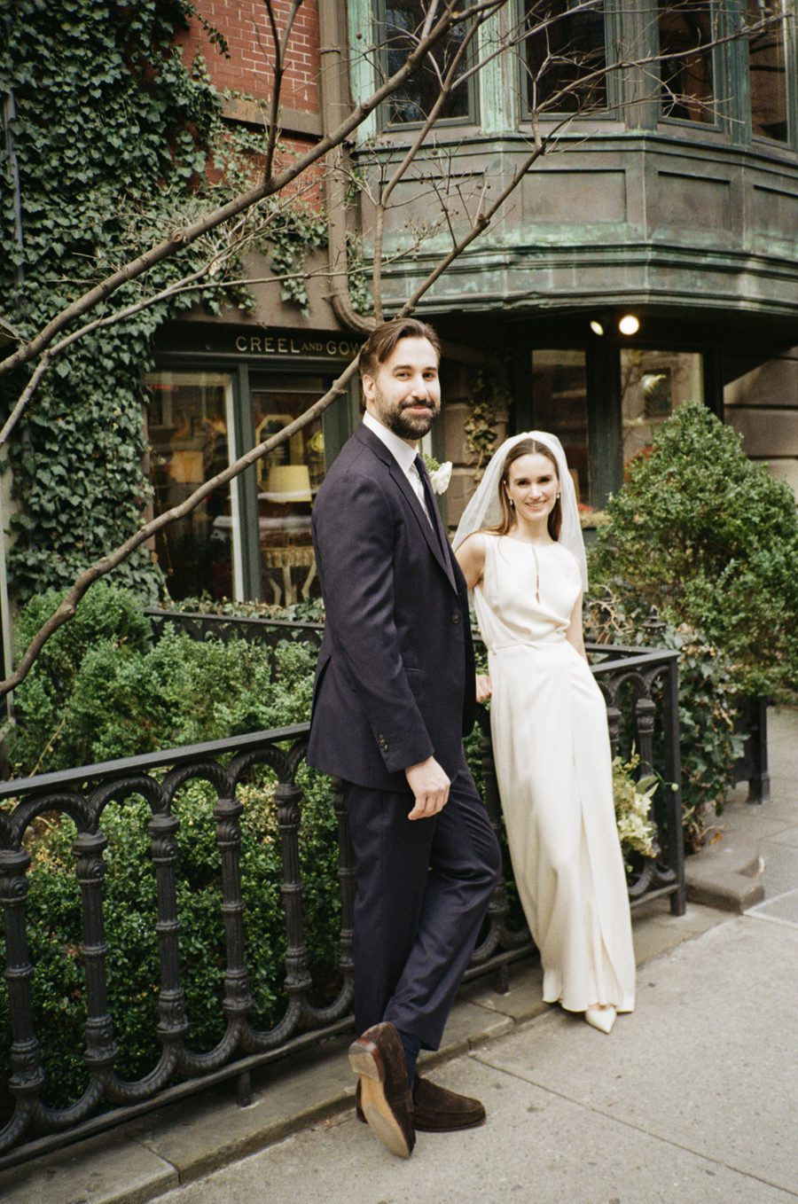 A bride in a white gown and veil stands beside a groom in a dark suit on a city sidewalk in front of a storefront.