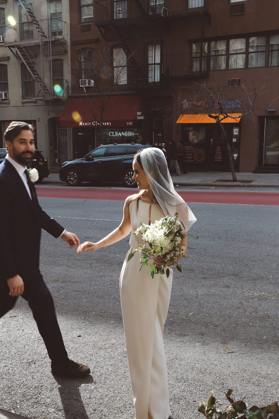 Bride in a white wedding dress and veil holding a bouquet, walking hand in hand with a man in a suit on a city street.