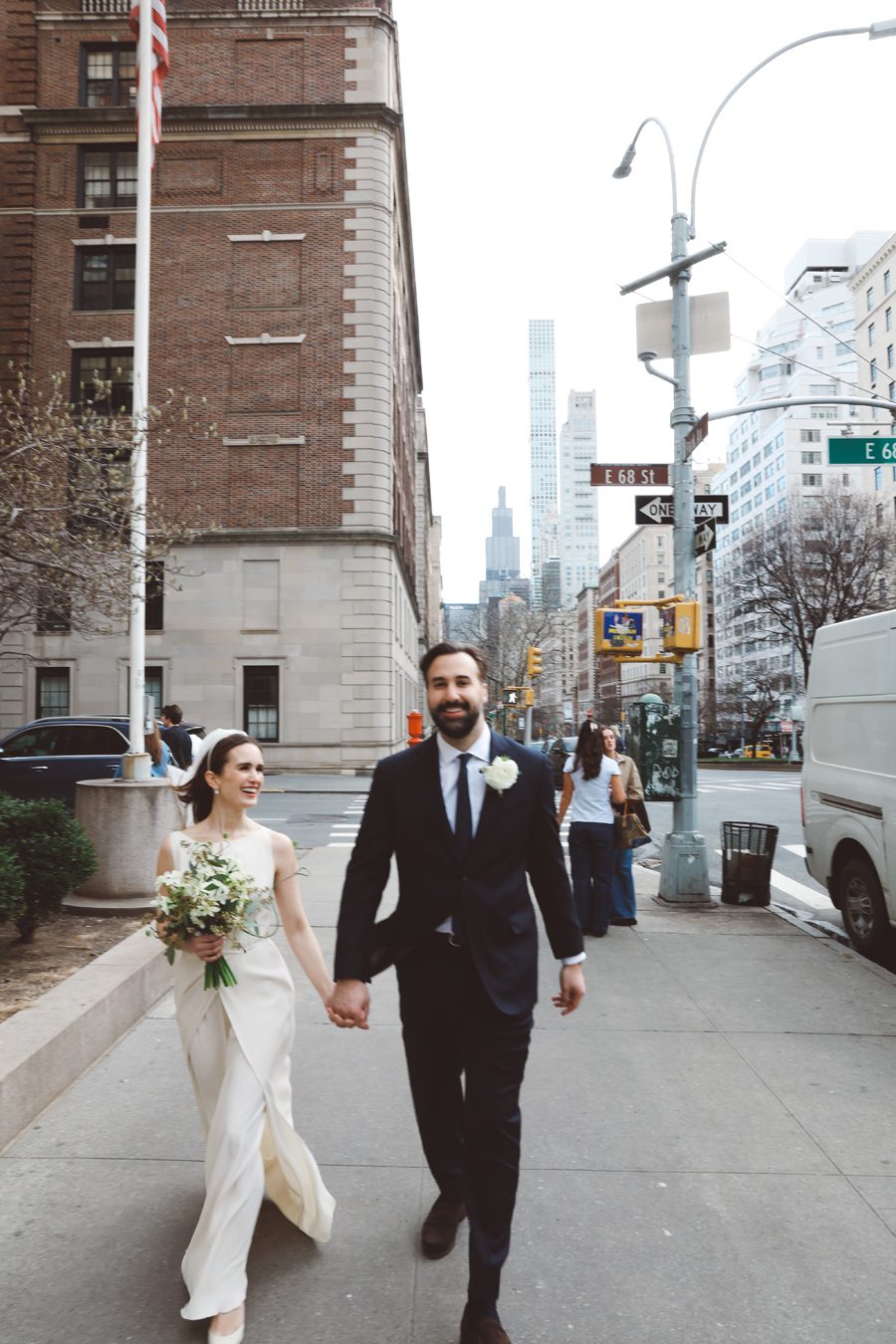 Bride in a white wedding dress and groom in a dark suit holding hands as they walk down a city sidewalk, bouquet in hand, with tall buildings in the background.