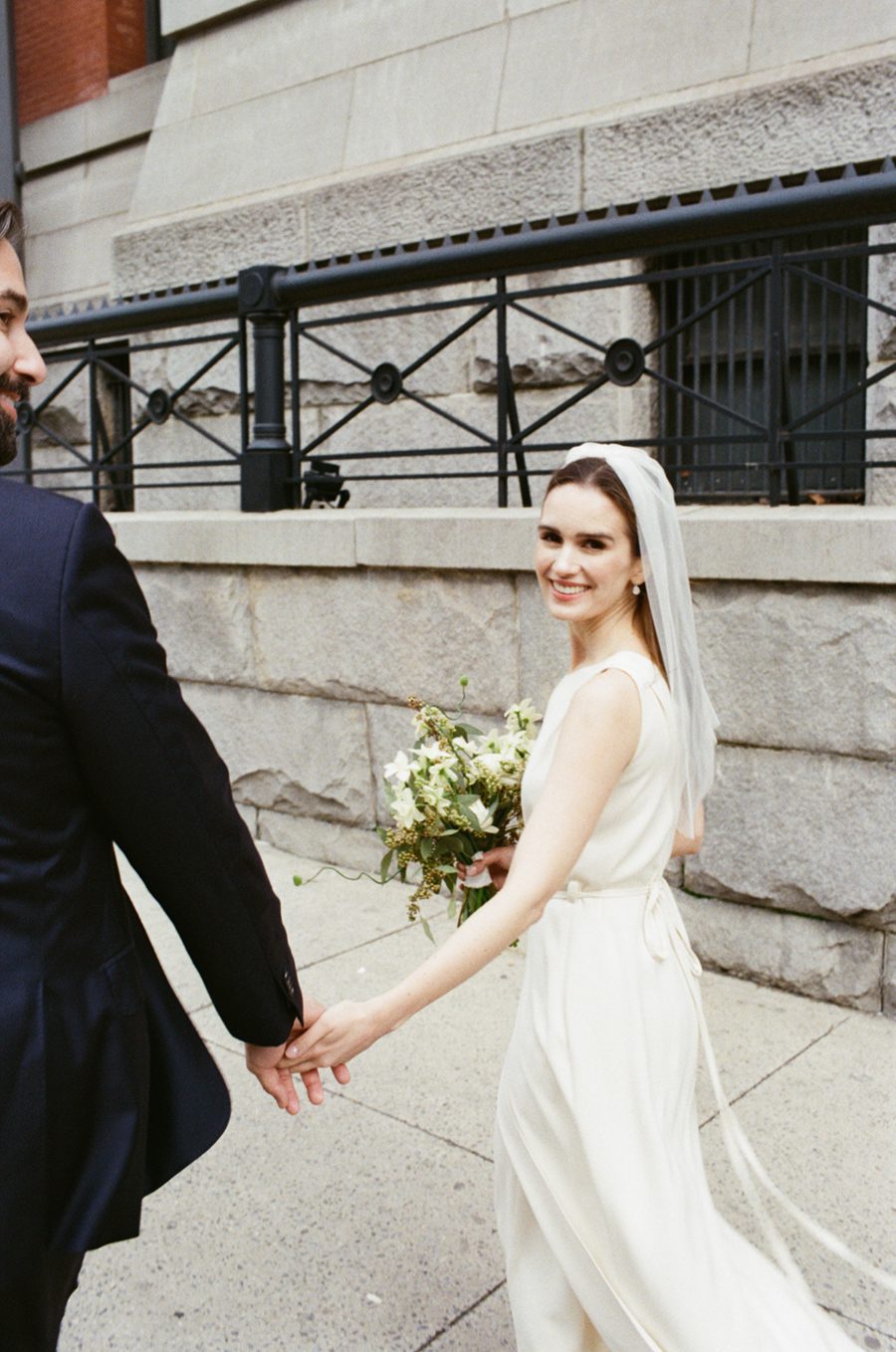 Bride in a white wedding dress and veil holds a bouquet while smiling and walking hand in hand with a groom in a dark suit outdoors, with a stone wall and black wrought-iron railing in the background.