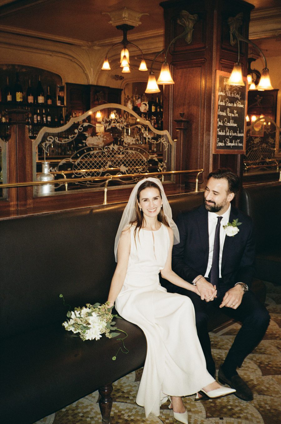 Bride in a white wedding dress with veil, seated beside groom in a dark suit, holding hands in a vintage bar setting with a bouquet beside her on the bench.