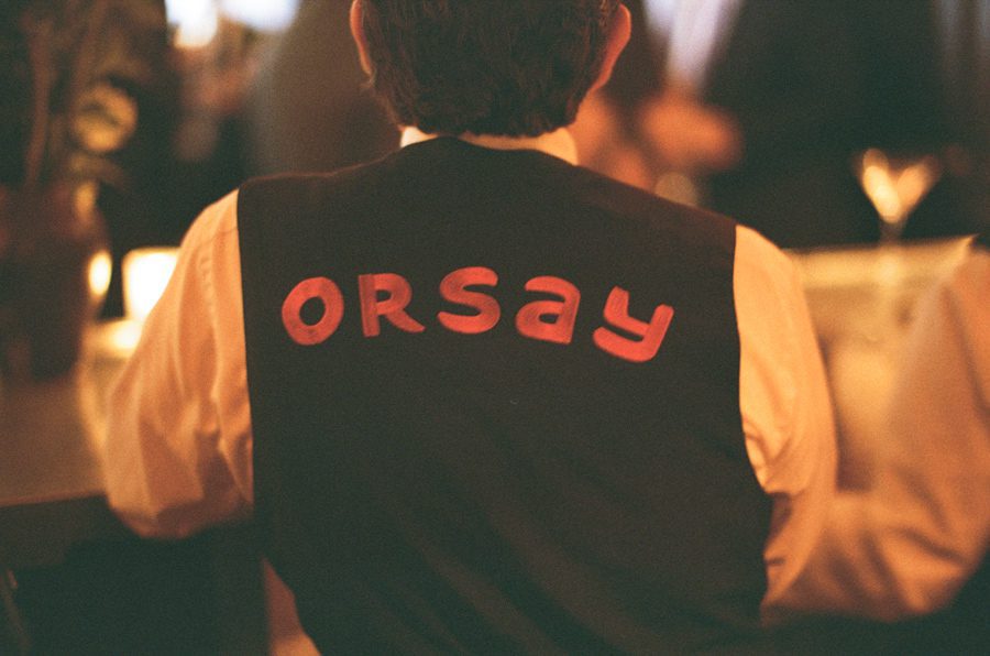 Back view of a person wearing a dark vest with red 'Orsay' lettering over a white shirt at a dimly lit event.