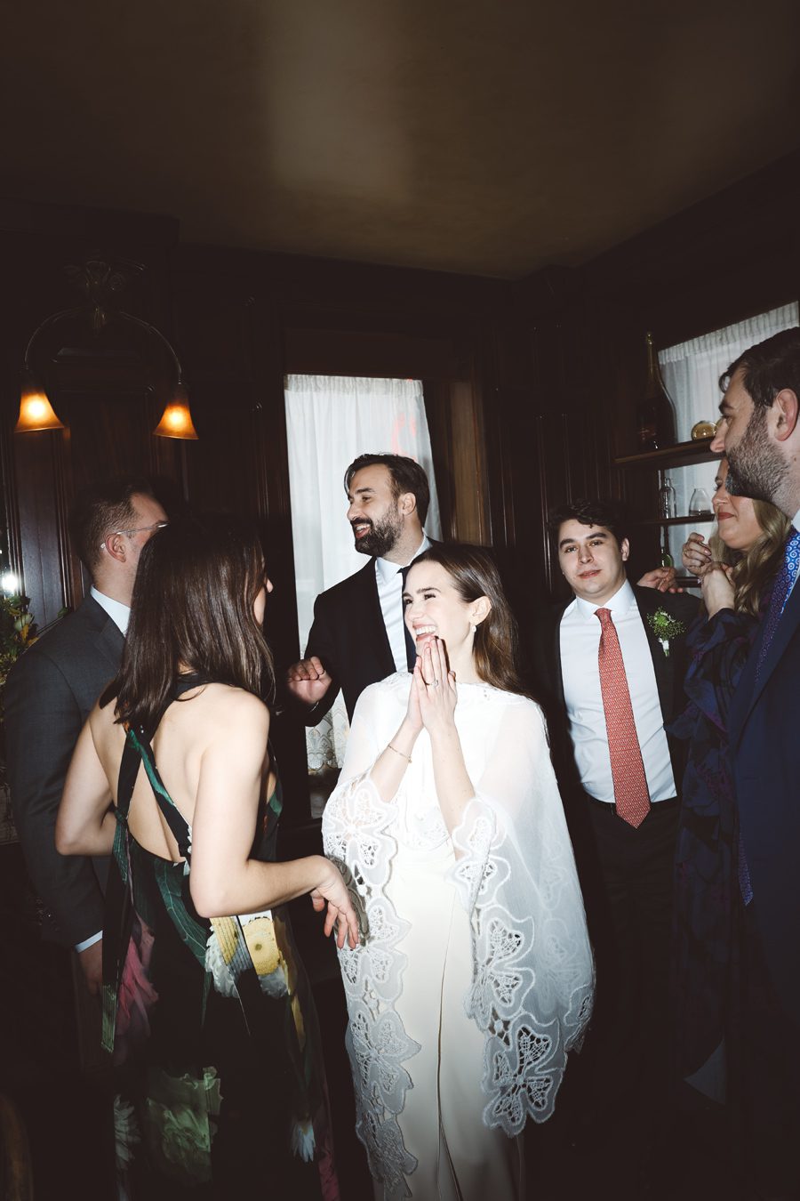 Bride in a white lace wedding dress laughs with friends at an indoor reception surrounded by warmly dressed guests.