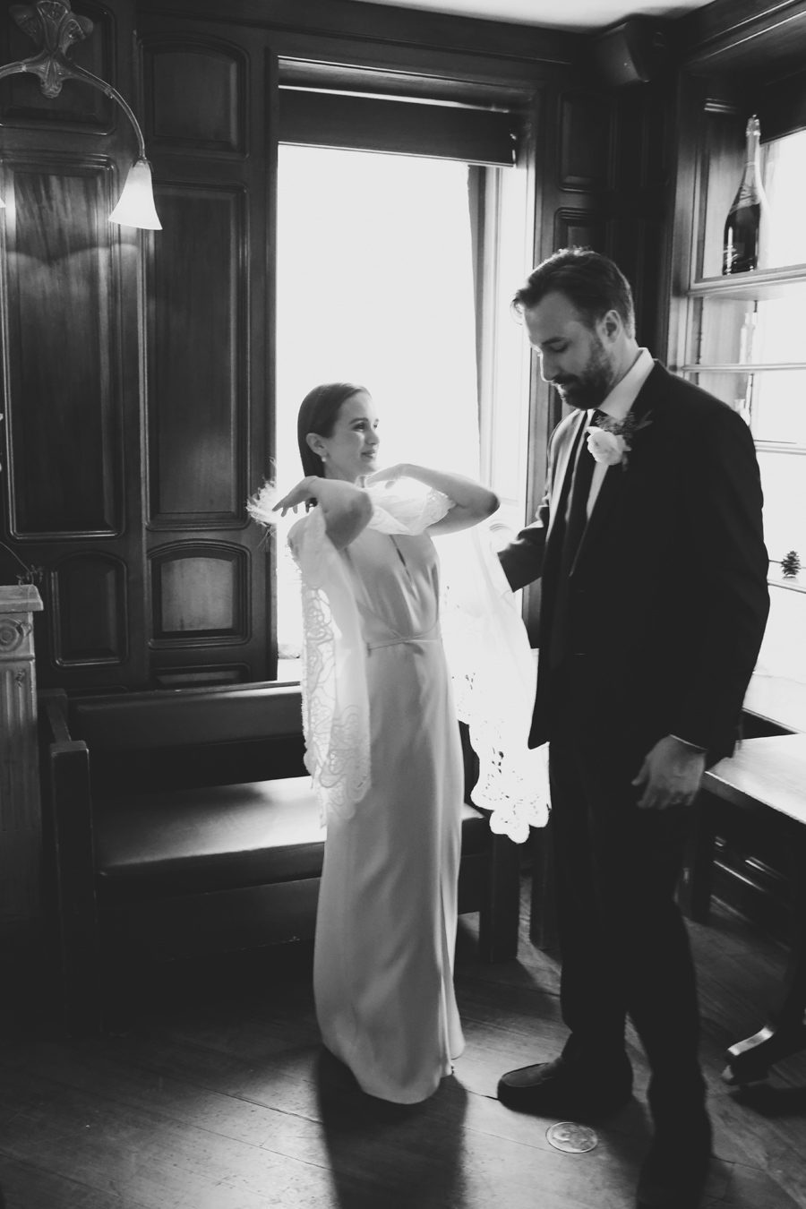 Bride adjusts her lace veil for her wedding as the groom in a dark suit looks on in a wood-paneled room (black and white).