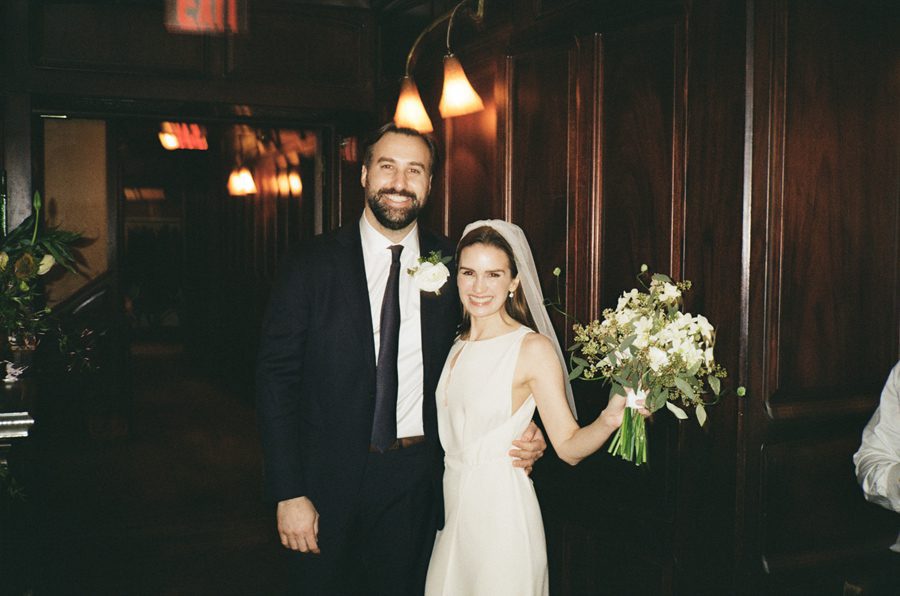 Smiling bride in a white wedding dress and veil stands beside a tall groom in a dark suit, holding a white bouquet as they pose in a wood-paneled hallway.