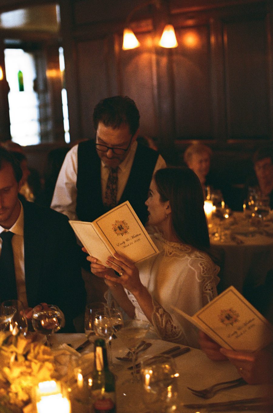 Bride wearing a white lace dress and a man in a suit read menu cards at a candlelit wedding reception.