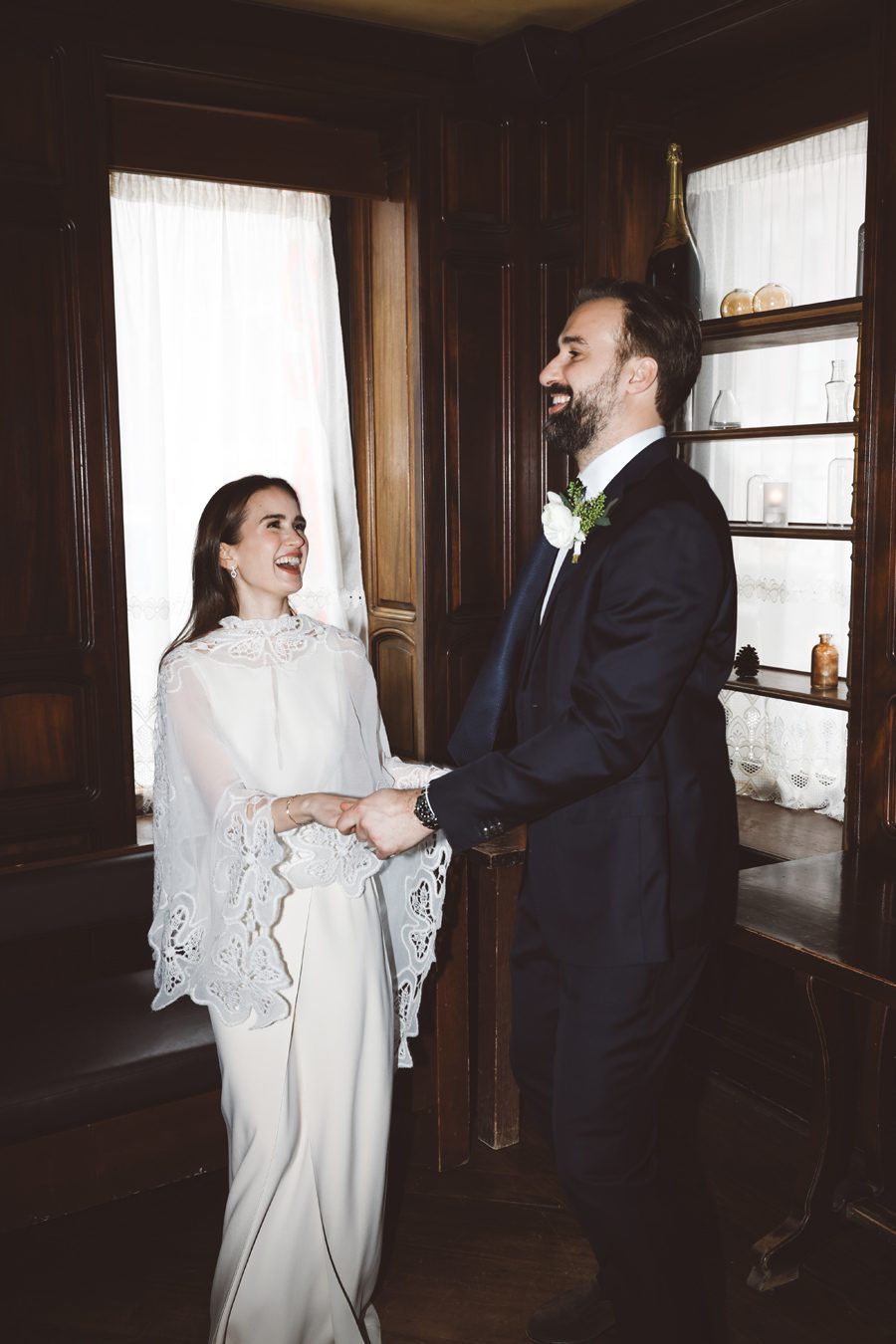 Bride in a white lace wedding dress and a bearded groom in a dark suit hold hands and laugh in a wood-paneled room.