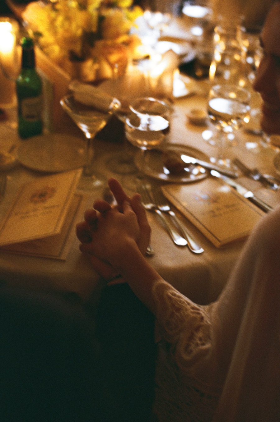 Couple holds hands across a candlelit dining table with formal place settings and glassware.