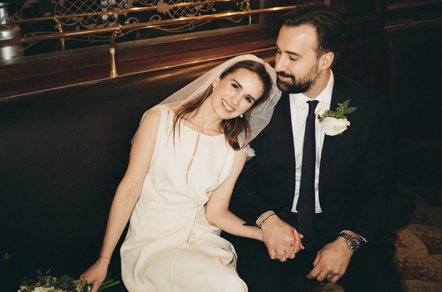 Bride wearing a white dress and veil leans her head toward a bearded groom in a dark suit as they hold hands on a bench at a wedding venue.