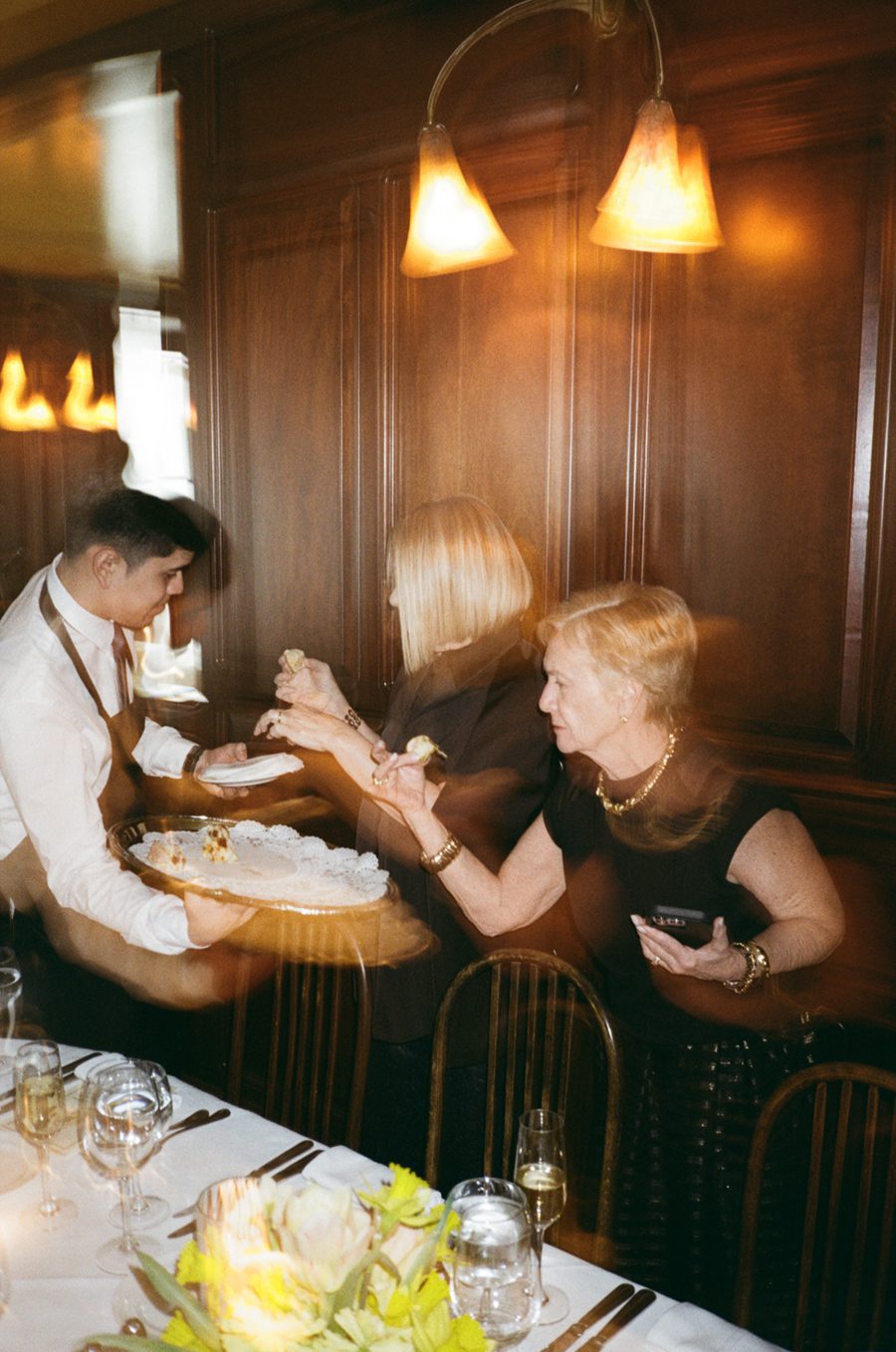 Waiter in a white shirt and apron serves cake to two women at a formal dinner beside a wood-paneled wall.