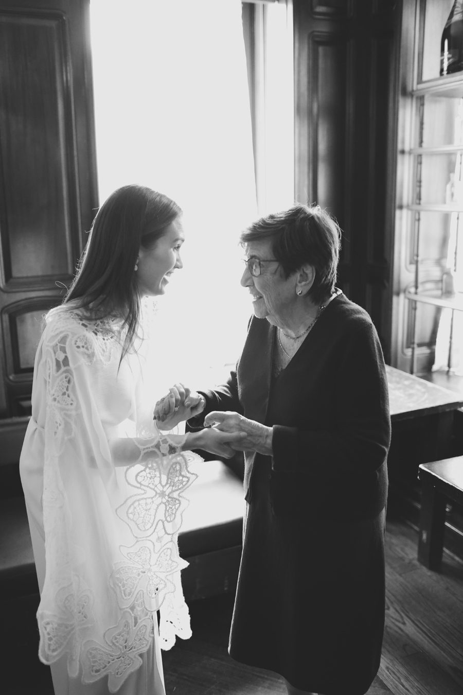 Bride in a lace wedding dress smiles as she holds hands with an elderly woman in a dark cardigan, sharing a joyful moment by a sunlit window.