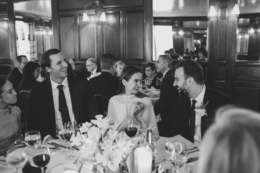Black-and-white wedding reception scene with three guests at a round table laughing and conversing, floral centerpiece in foreground.