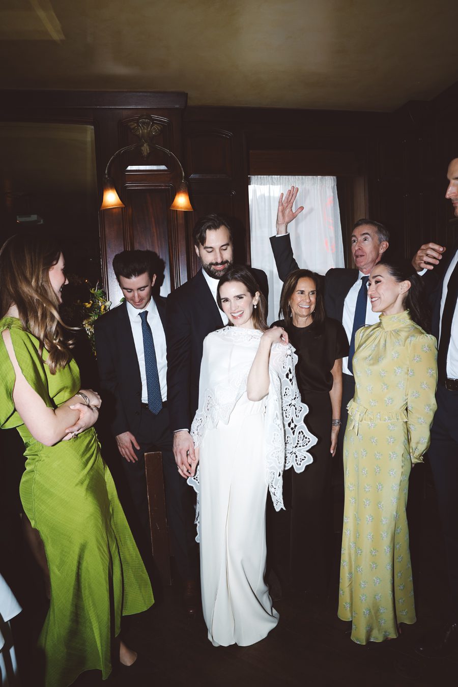 Group of well-dressed guests posing in a dimly lit room at a wedding reception, with the bride in white center stage.