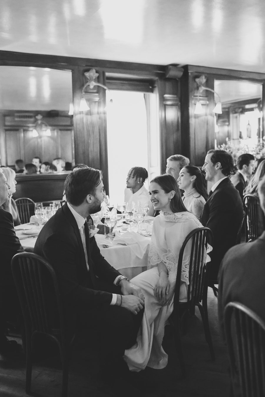 Couple in formal attire sitting at a banquet table, smiling and talking during a black-and-white wedding reception.