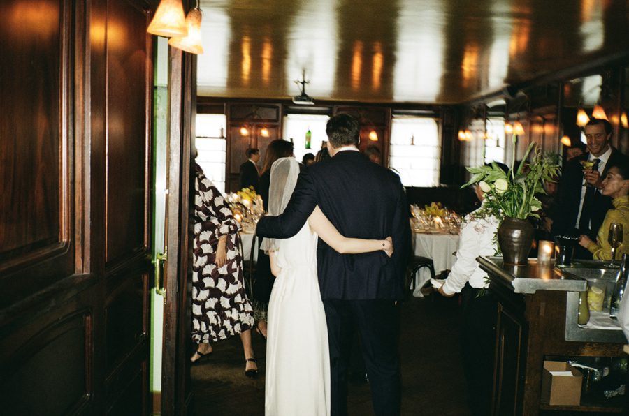 Bride and groom embrace while dancing at a wedding reception, surrounded by guests in a warmly lit room with a bar on the side.
