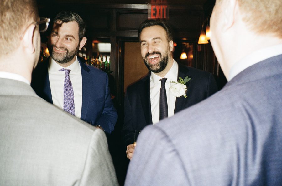 Two bearded men in suits share a laugh at a formal event, one with a white boutonniere on his lapel