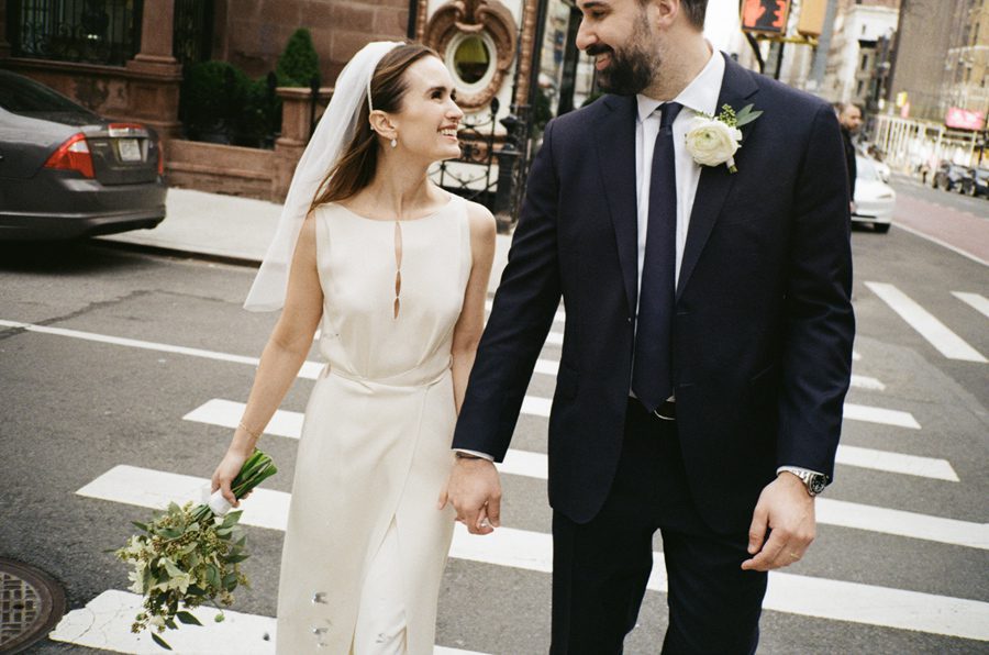Bride in a white sleeveless gown with a veil walks hand in hand with a groom in a navy suit on a city street, crosswalk behind them.