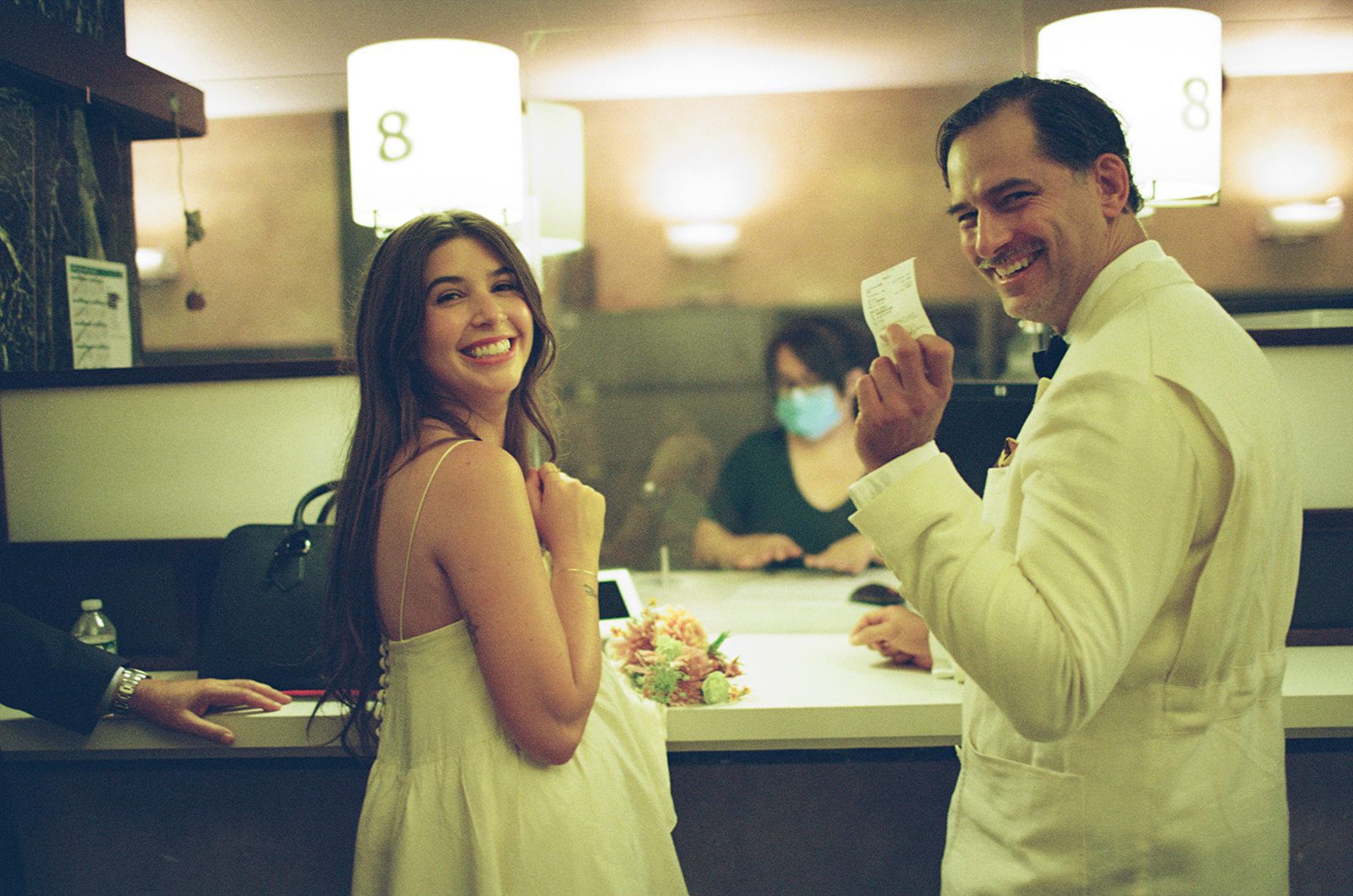 Smiling couple at a reception counter: woman in white dress, man in a white suit holds a receipt and smiles at the camera with flowers on the counter behind them.